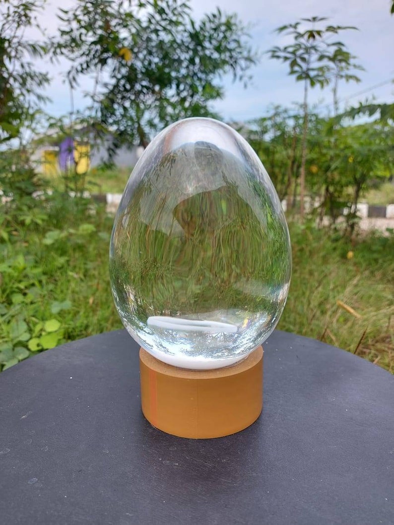 Pode incluir: Um ornamento de vidro transparente em forma de ovo com uma base dourada. O ornamento est&aacute; sobre uma superf&iacute;cie preta.