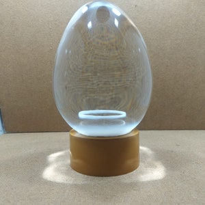 Pode incluir: Um ornamento de vidro transparente em forma de ovo com uma base dourada. O ornamento tem um anel branco no interior.
