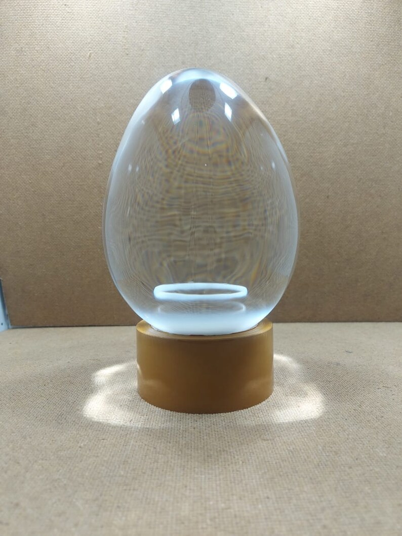 Pode incluir: Um ornamento de vidro transparente em forma de ovo com uma base dourada. O ornamento tem um anel branco no interior.