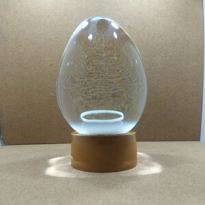 Pode incluir: Um domo de vidro transparente em forma de ovo com um anel branco no interior, sentado em uma base marrom.