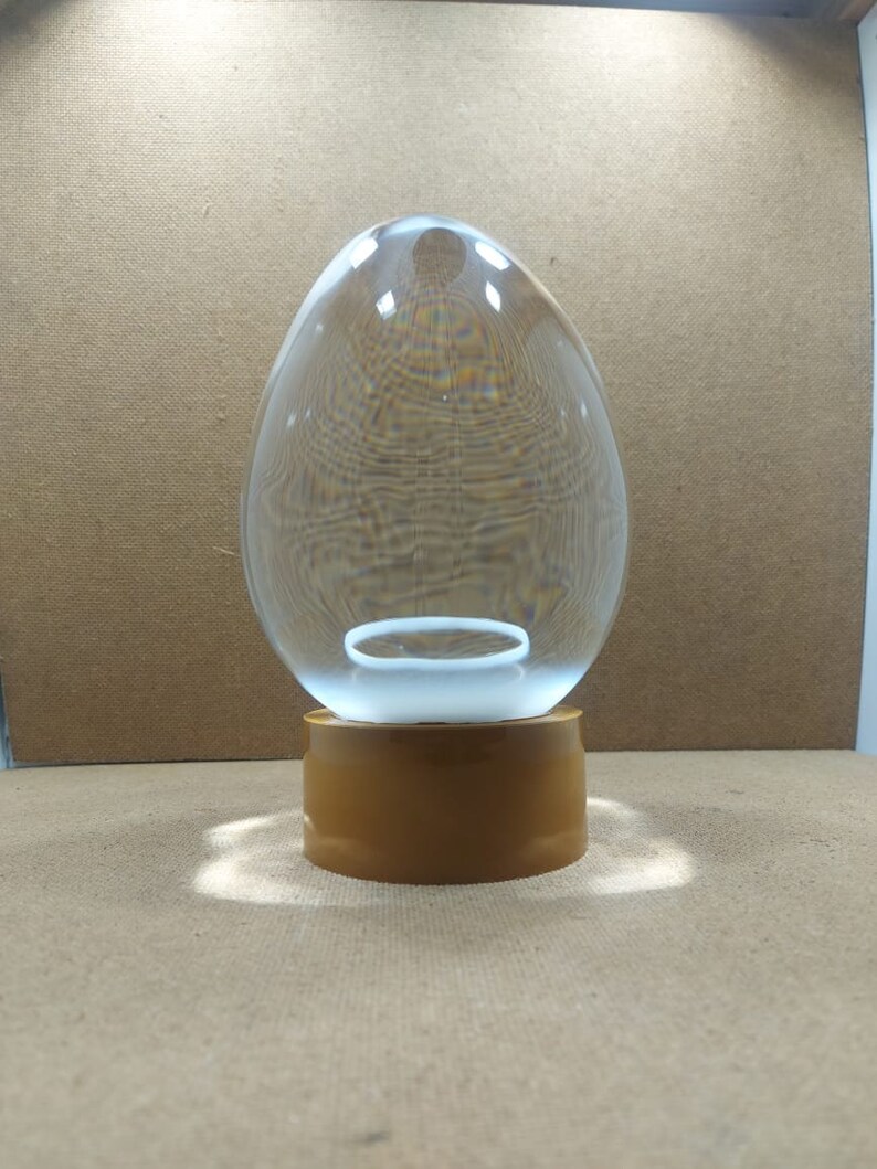 Pode incluir: Um domo de vidro transparente em forma de ovo com um anel branco no interior, sentado em uma base marrom.