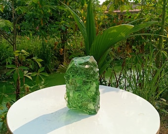 May include: A large, green, glass rock with an irregular shape sits on a white table. The rock is made of clear glass with a green tint and has a rough, textured surface.
