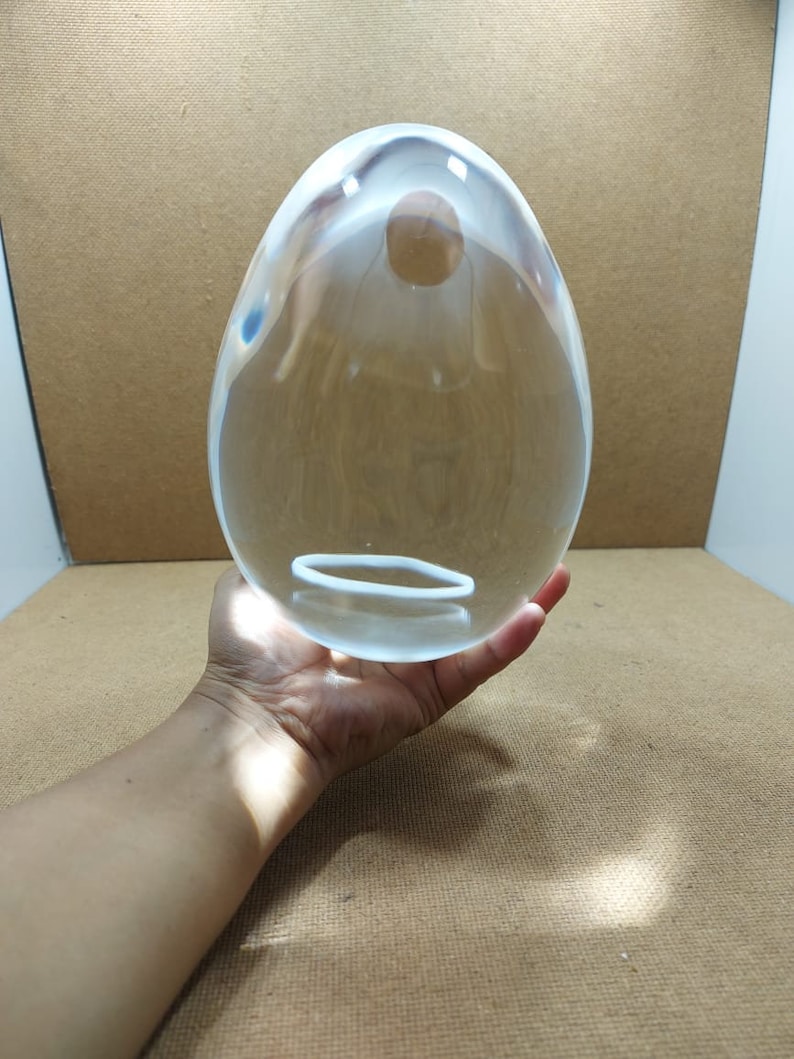 Pode incluir: Um ovo grande de vidro transparente com um anel branco no meio. O ovo est&aacute; sendo segurado na m&atilde;o de uma pessoa.