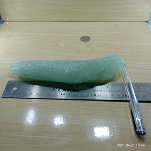 May include: A light green sea glass piece, approximately 20 inches long, with a rough, textured surface. The sea glass is lying on a wooden surface next to a ruler and a pen.