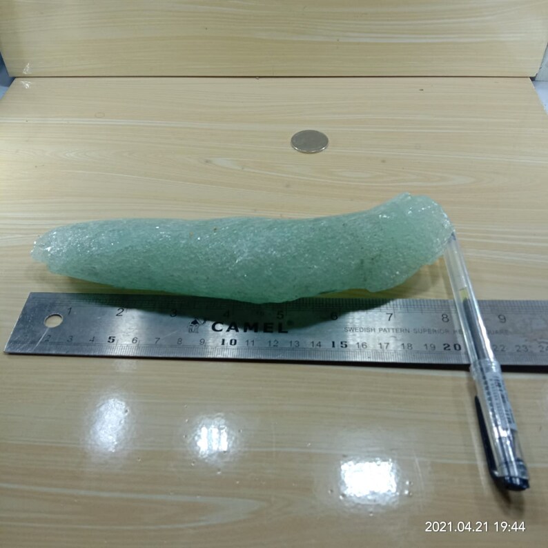 May include: A light green sea glass piece, approximately 20 inches long, with a rough, textured surface. The sea glass is lying on a wooden surface next to a ruler and a pen.