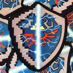 May include: A blue and red embroidered patch with a pixelated design of a shield with lightning bolts and a sword. The shield is split in half, with the left side showing a pixelated design and the right side showing a stylized winged design.