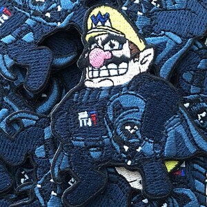 May include: A blue embroidered patch of a cartoon character with a yellow hat and a mustache. The character is wearing a blue uniform with a red and white badge. The patch is surrounded by other blue patches.