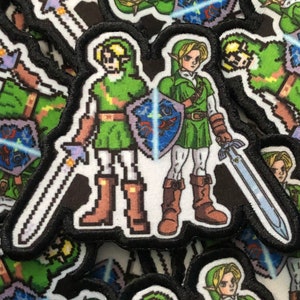 May include: Embroidered patch featuring a pixelated image of a character in a green tunic, brown boots, and a sword. The character is holding a shield with a blue and white design. The patch has a black border.