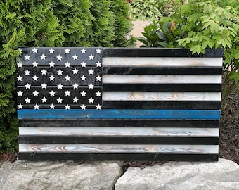 Op de afbeelding: Een Amerikaanse vlag van hout met een blauwe lijn die horizontaal over het midden van de vlag loopt. De vlag is zwart-wit geverfd met witte sterren op het blauwe veld.