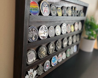 May include: A dark brown wooden display case with multiple rows of challenge coins. The coins feature various designs, including a rainbow police badge, a red ribbon, and a maple leaf.