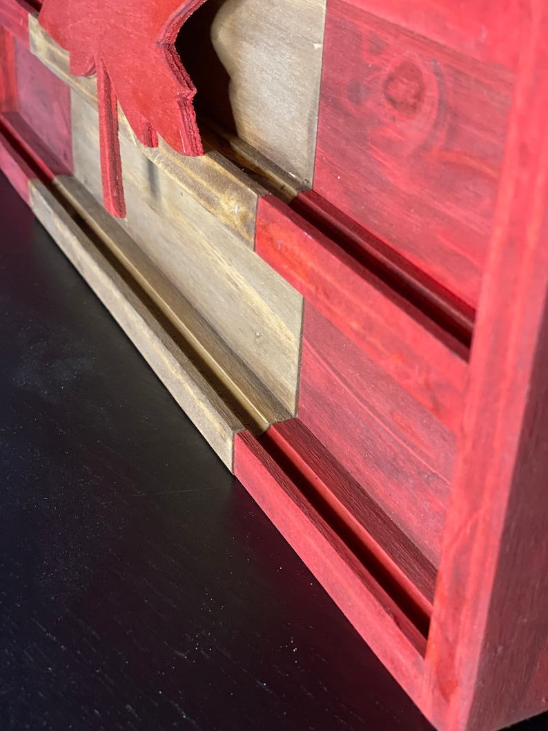 Op de afbeelding: Een houten Canadese vlag met een rood esdoornblad, geschilderd in rood, wit en bruin. De vlag is gemonteerd op een donker houten oppervlak.