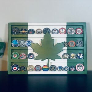May include: A wooden shadow box display with a green, white, and red Canadian flag design. The shadow box has multiple rows of round metal challenge coins with various designs. A green maple leaf cutout is centered on the display.