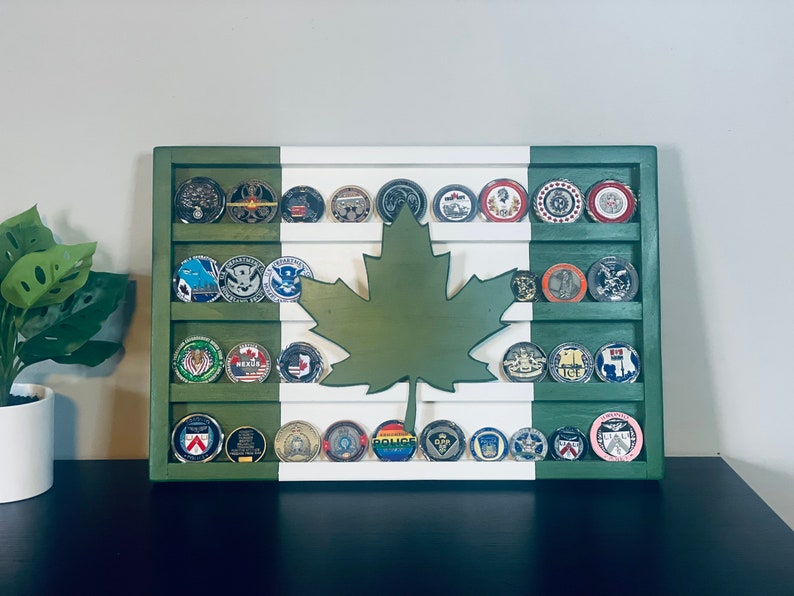 May include: A wooden shadow box display with a green, white, and red Canadian flag design. The shadow box has multiple rows of round metal challenge coins with various designs. A green maple leaf cutout is centered on the display.