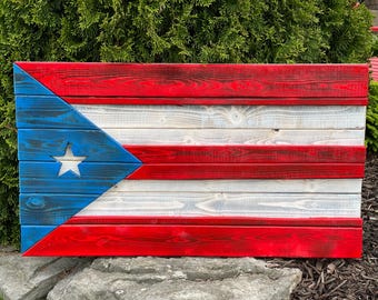 Op de afbeelding: Een houten wanddecoratie met de vlag van Puerto Rico. Het ontwerp omvat afwisselende rode en witte horizontale strepen, een blauwe driehoek met een witte ster en is gemaakt van gebeitst hout.