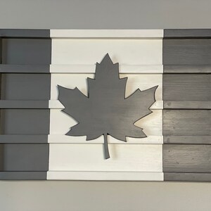 May include: A wooden wall decor in the shape of the Canadian flag. The flag is painted white, gray, and black. A large black maple leaf is in the center of the white section. The flag is divided into three sections, with the gray sections on the left and right and the white section in the middle.