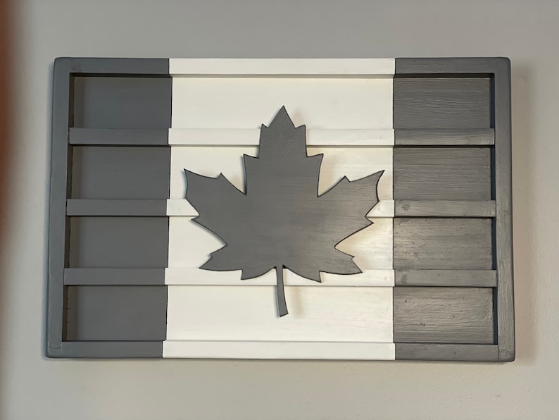 May include: A wooden wall decor in the shape of the Canadian flag. The flag is painted white, gray, and black. A large black maple leaf is in the center of the white section. The flag is divided into three sections, with the gray sections on the left and right and the white section in the middle.