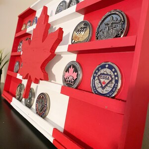 May include: A wooden Canadian flag display with shelves for displaying challenge coins. The flag is painted red and white with a maple leaf in the center. The shelves are filled with various challenge coins with different designs.