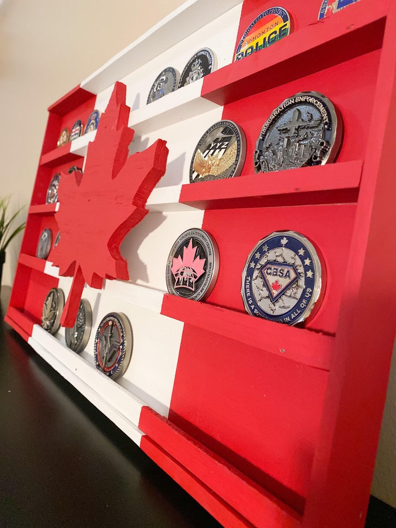 May include: A wooden Canadian flag display with shelves for displaying challenge coins. The flag is painted red and white with a maple leaf in the center. The shelves are filled with various challenge coins with different designs.