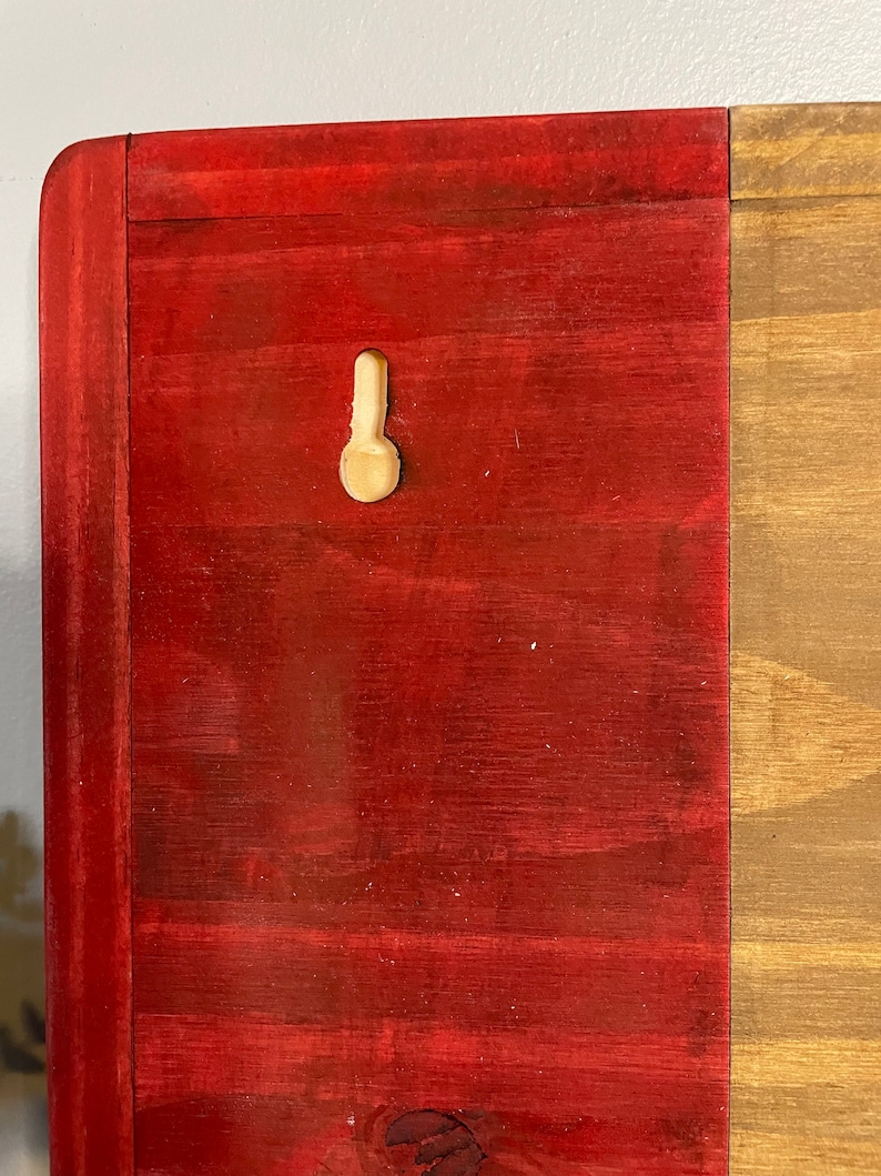Op de afbeelding: Een houten bord met een rode en bruine afwerking. Het bord heeft een kleine, witte plastic haak aan de rode kant.