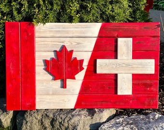 Op de afbeelding: Een houten wanddecoratie met de vlaggen van Canada en Zwitserland. De Canadese vlag heeft een rood esdoornblad op een witte achtergrond en de Zwitserse vlag heeft een wit kruis op een rode achtergrond. De vlaggen zijn gemaakt van geschilderd hout.