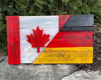 Op de afbeelding: Houten wandkunst met een combinatie van de Canadese en Duitse vlag. De Canadese kant heeft een rood esdoornblad op een witte achtergrond, terwijl de Duitse kant zwarte, rode en gele strepen heeft. Het kunstwerk is gemaakt van geschilderd hout.