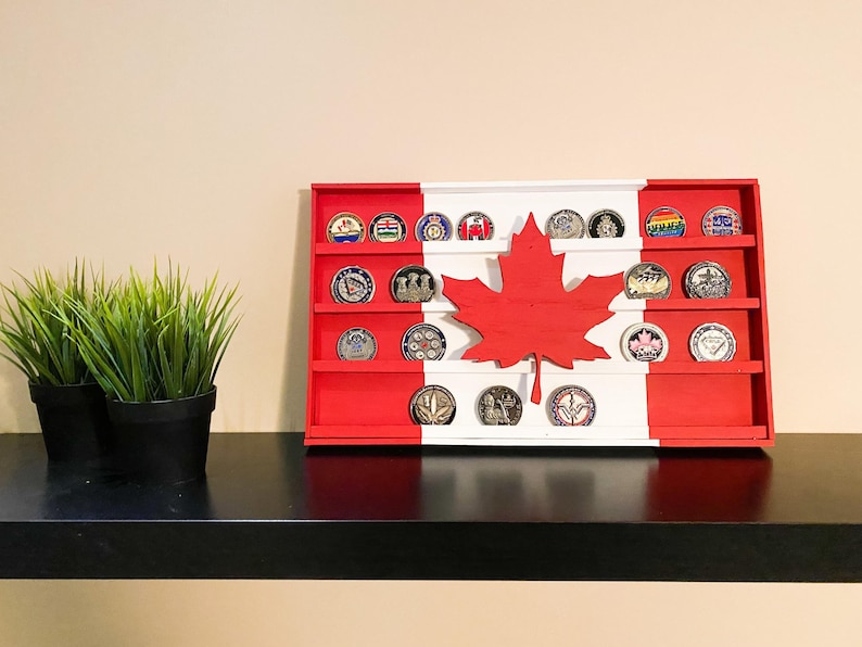 Op de afbeelding: Een houten displaykast in de vorm van de Canadese vlag met rode en witte strepen en een esdoornblad in het midden. De kast heeft vijf planken met ronde muntgleuven. De kast staat op een zwarte plank.