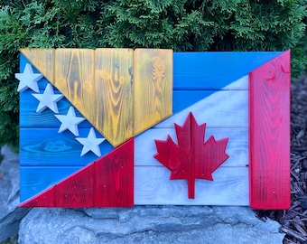 Op de afbeelding: Een houten bord met de vlaggen van de Verenigde Staten en Canada erop geschilderd. De Amerikaanse vlag bevindt zich aan de linkerkant van het bord en de Canadese vlag aan de rechterkant. De vlaggen zijn geschilderd in hun traditionele kleuren rood, wit en blauw.