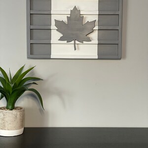 May include: A wooden wall decor in the shape of the Canadian flag. The flag is painted in white and gray with a gray maple leaf in the center.