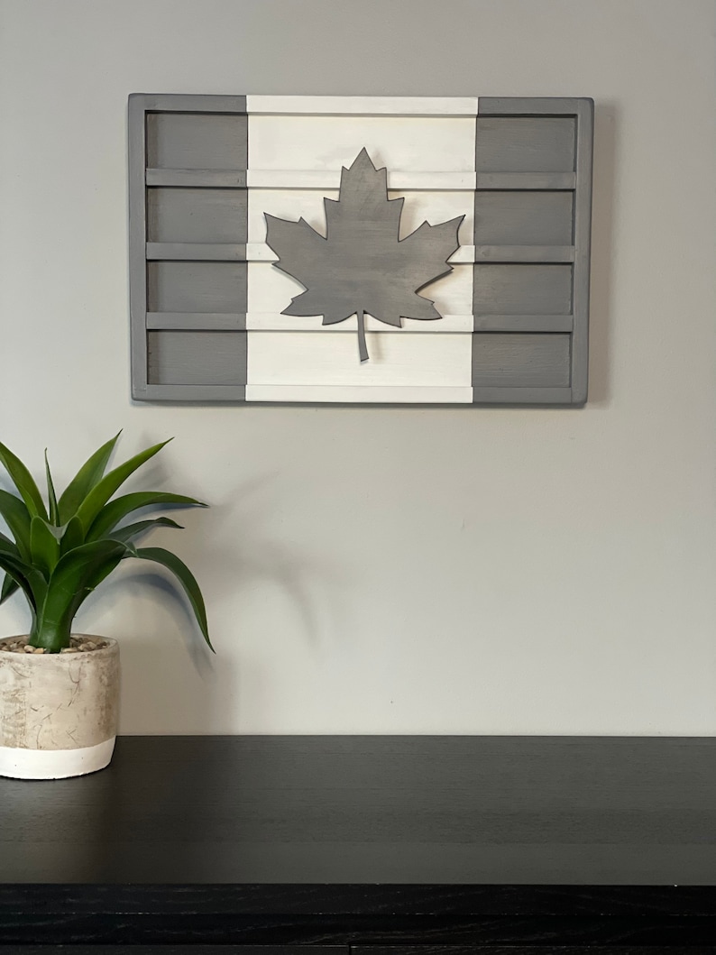 May include: A wooden wall decor in the shape of the Canadian flag. The flag is painted in white and gray with a gray maple leaf in the center.