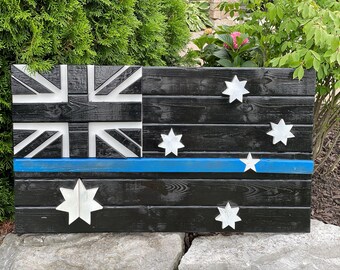 Op de afbeelding: Een houten Australische vlag met een blauwe lijn die horizontaal door het midden loopt. De vlag is zwart geverfd met witte sterren en de Union Jack in de linkerbovenhoek.