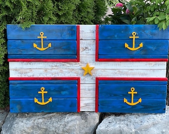 Op de afbeelding: Een houten bord geschilderd om eruit te zien als een vlag met een blauwe achtergrond, een wit kruis en vier gele ankers. Het kruis heeft een rode rand en een gele ster in het midden.
