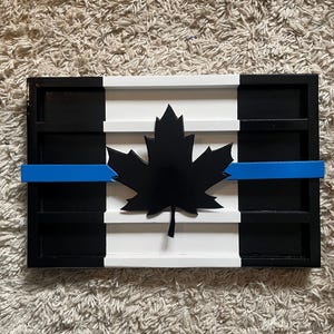 Op de afbeelding: Een houten Canadese vlag met een blauwe lijn in het midden, die steun voor rechtshandhaving symboliseert. De vlag is zwart, wit en blauw geverfd en heeft een zwart esdoornblad in het midden.
