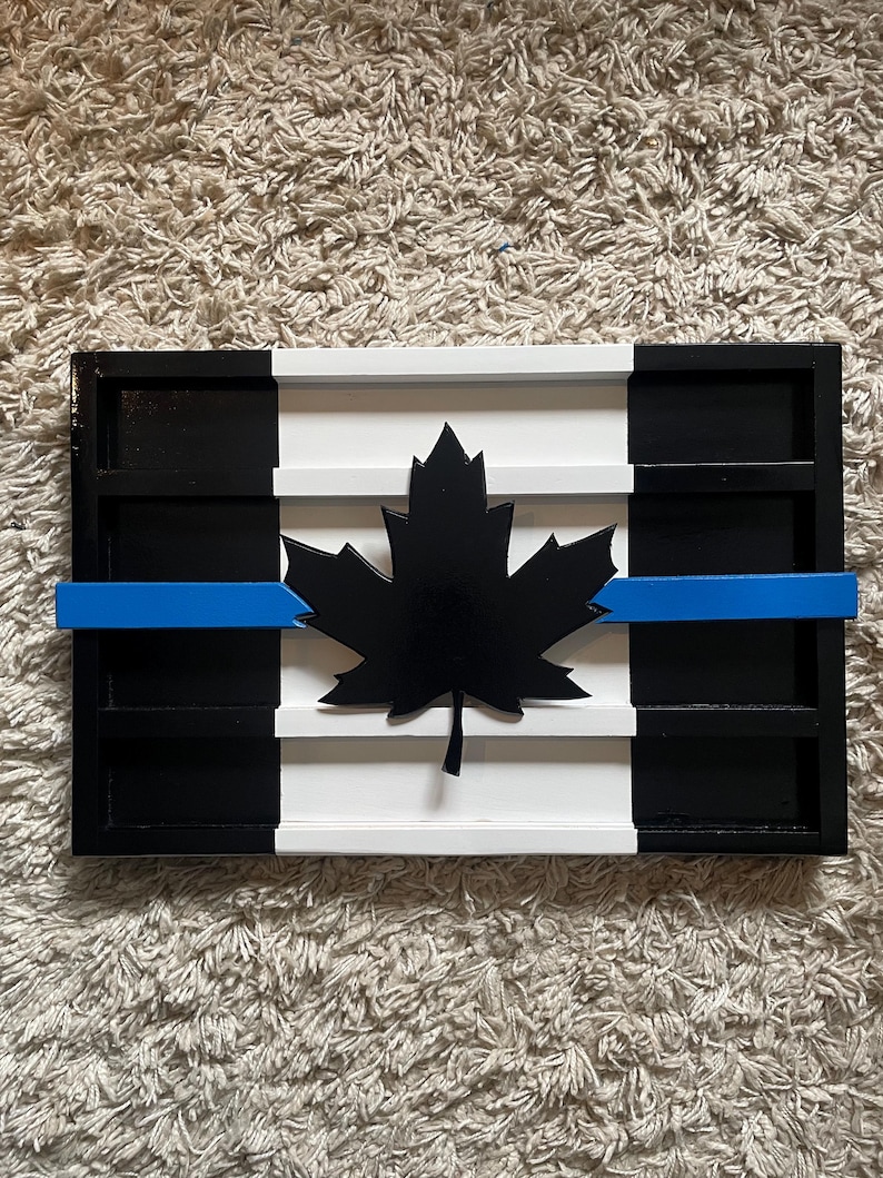 Op de afbeelding: Een houten Canadese vlag met een blauwe lijn in het midden, die steun voor rechtshandhaving symboliseert. De vlag is zwart, wit en blauw geverfd en heeft een zwart esdoornblad in het midden.
