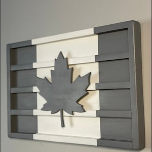 Op de afbeelding: Een houten wanddecoratie in de vorm van de Canadese vlag. De vlag is wit en grijs geschilderd met een groot zwart esdoornblad in het midden.