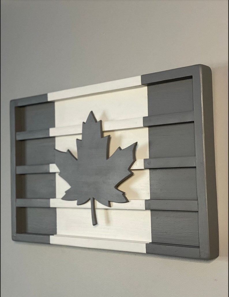 Op de afbeelding: Een houten wanddecoratie in de vorm van de Canadese vlag. De vlag is wit en grijs geschilderd met een groot zwart esdoornblad in het midden.
