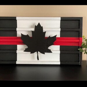 May include: A wooden sign with a black and white Canadian flag design. The flag has a red stripe across the middle and a black maple leaf in the center. The sign is made of wood and has a distressed look.