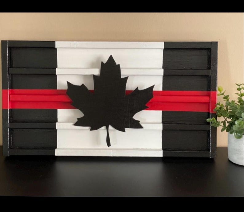 May include: A wooden sign with a black and white Canadian flag design. The flag has a red stripe across the middle and a black maple leaf in the center. The sign is made of wood and has a distressed look.