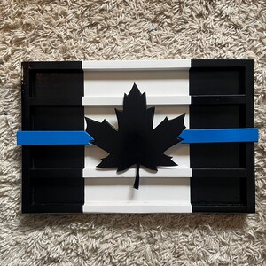 May include: A wooden Canadian flag with a blue line across the middle, representing support for law enforcement. The flag is painted black, white, and blue, and features a black maple leaf in the center.