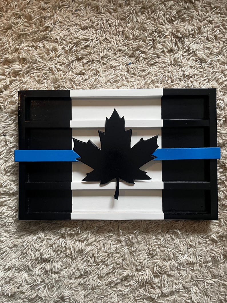 May include: A wooden Canadian flag with a blue line across the middle, representing support for law enforcement. The flag is painted black, white, and blue, and features a black maple leaf in the center.