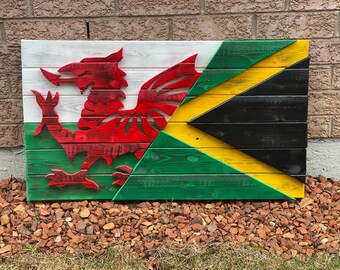 Op de afbeelding: Een houten bord met de vlaggen van Wales en Jamaica erop geschilderd. De Welshe vlag heeft een rode draak op een witte achtergrond met een groene rand. De Jamaicaanse vlag heeft een diagonaal geel kruis op een zwarte achtergrond met groene en gele driehoeken.