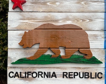 Op de afbeelding: Een houten bord met de vlag van de Republiek Californië. De vlag heeft een rode ster, een bruine beer, groen gras en de woorden "CALIFORNIA REPUBLIC" in zwarte letters. Het bord is wit, rood en blauw geverfd.