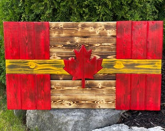 Op de afbeelding: Houten Canadese vlag met een rode esdoorn en rode vierkanten op een natuurlijke houten achtergrond. De vlag heeft een gele streep en is gemaakt van gebeitst hout. Het rustieke ontwerp is geschikt voor woondecoratie.