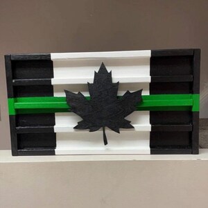 May include: A wooden sign with a black and white striped background and a green stripe across the middle. A black maple leaf is centered on the sign.