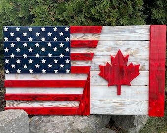 Op de afbeelding: Een houten bord met de Amerikaanse en Canadese vlag. De Amerikaanse vlag bevindt zich aan de linkerkant van het bord en de Canadese vlag aan de rechterkant. De vlaggen zijn in hun respectievelijke kleuren rood, wit en blauw geschilderd.