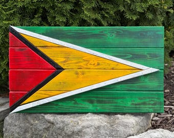 Op de afbeelding: Houten wandkunst met de vlag van Guyana. Het ontwerp omvat een rode driehoek met een zwarte rand, een gele driehoek, een witte rand en een groene achtergrond. De kunst is gemaakt van houten planken.