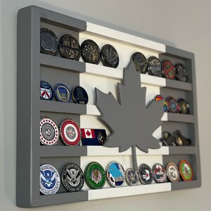May include: A gray and white wooden shadow box with a gray maple leaf cutout. The shadow box has five shelves with various challenge coins displayed on each shelf. The coins feature different logos and designs, including the Department of Homeland Security logo.