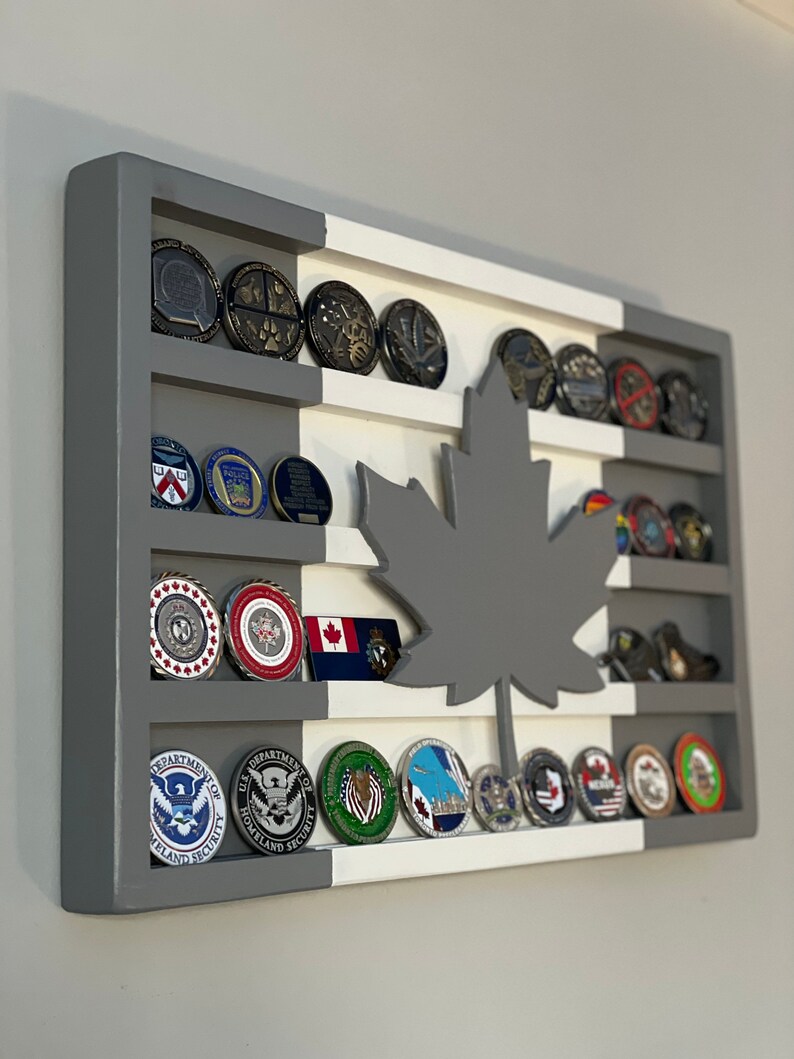 May include: A gray and white wooden shadow box with a gray maple leaf cutout. The shadow box has five shelves with various challenge coins displayed on each shelf. The coins feature different logos and designs, including the Department of Homeland Security logo.