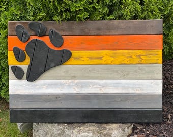 Op de afbeelding: Een houten bord met een berenpootafdruk. Het bord is gemaakt van horizontale planken, geschilderd in verschillende kleuren, waaronder bruin, oranje, geel, grijs, wit en zwart. De berenpoot is zwart en driedimensionaal.