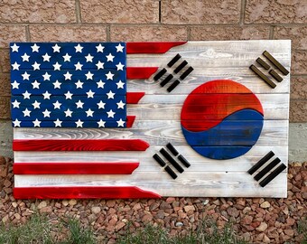 Op de afbeelding: Een houten bord met de Amerikaanse en Zuid-Koreaanse vlag. De Amerikaanse vlag bevindt zich aan de linkerkant en de Zuid-Koreaanse vlag aan de rechterkant. De vlaggen zijn geschilderd op houten planken en hebben een verweerde uitstraling.
