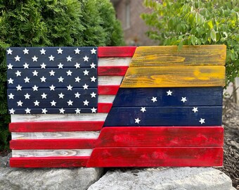 Op de afbeelding: Een houten bord in de vorm van een vlag, geschilderd om eruit te zien als de Amerikaanse vlag aan de linkerkant en de Venezolaanse vlag aan de rechterkant. Het bord is gemaakt van gerecycled hout en heeft een rustieke uitstraling.